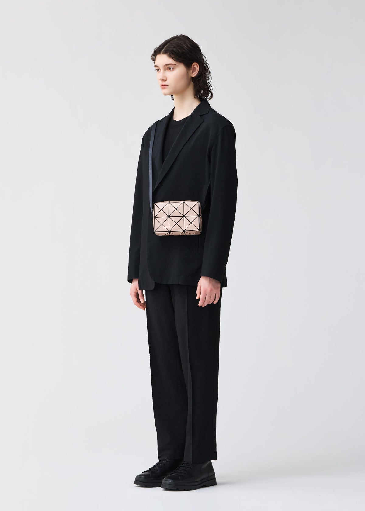A person with long brown hair, dressed in a loose black suit and shirt, holds the BAO BAO ISSEY MIYAKE CUBOID—a small geometric beige minimalist bag with adjustable strap. The background is plain and light-colored. view 7 of 8