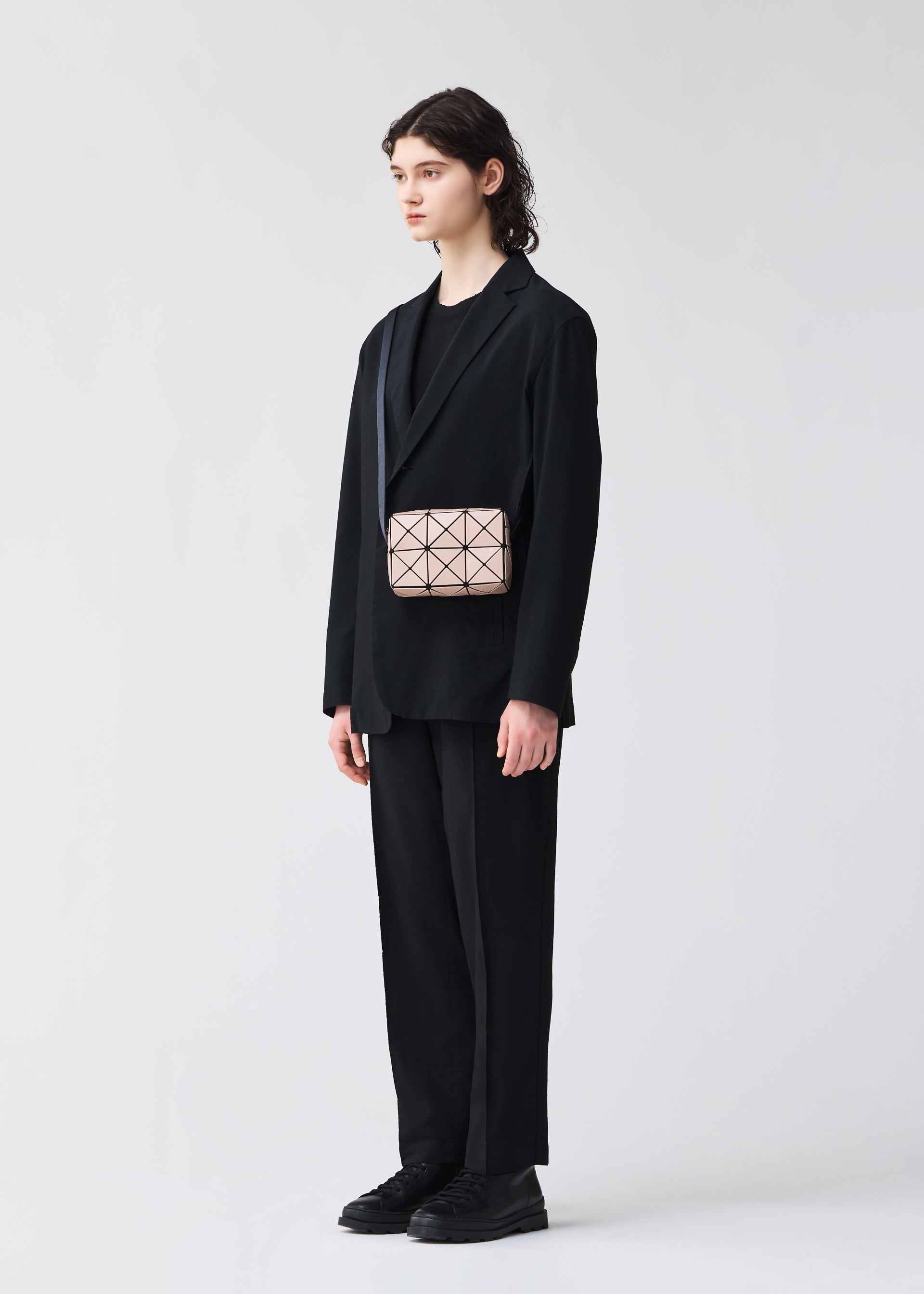 Zoomed view A person with long brown hair, dressed in a loose black suit and shirt, holds the BAO BAO ISSEY MIYAKE CUBOID—a small geometric beige minimalist bag with adjustable strap. The background is plain and light-colored.