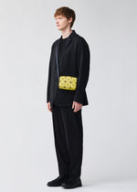 A person stands against a plain background wearing a loose black suit with a black shirt and shoes, accessorized with the BAO BAO ISSEY MIYAKE CUBOID—a small yellow geometric-patterned shoulder bag with an adjustable strap.