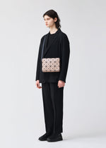 A person with long dark hair in a black suit, T-shirt, and shoes holds the BAO BAO ISSEY MIYAKE CUBOID—a beige minimalist bag with geometric patterns and an adjustable strap—against a plain light background.