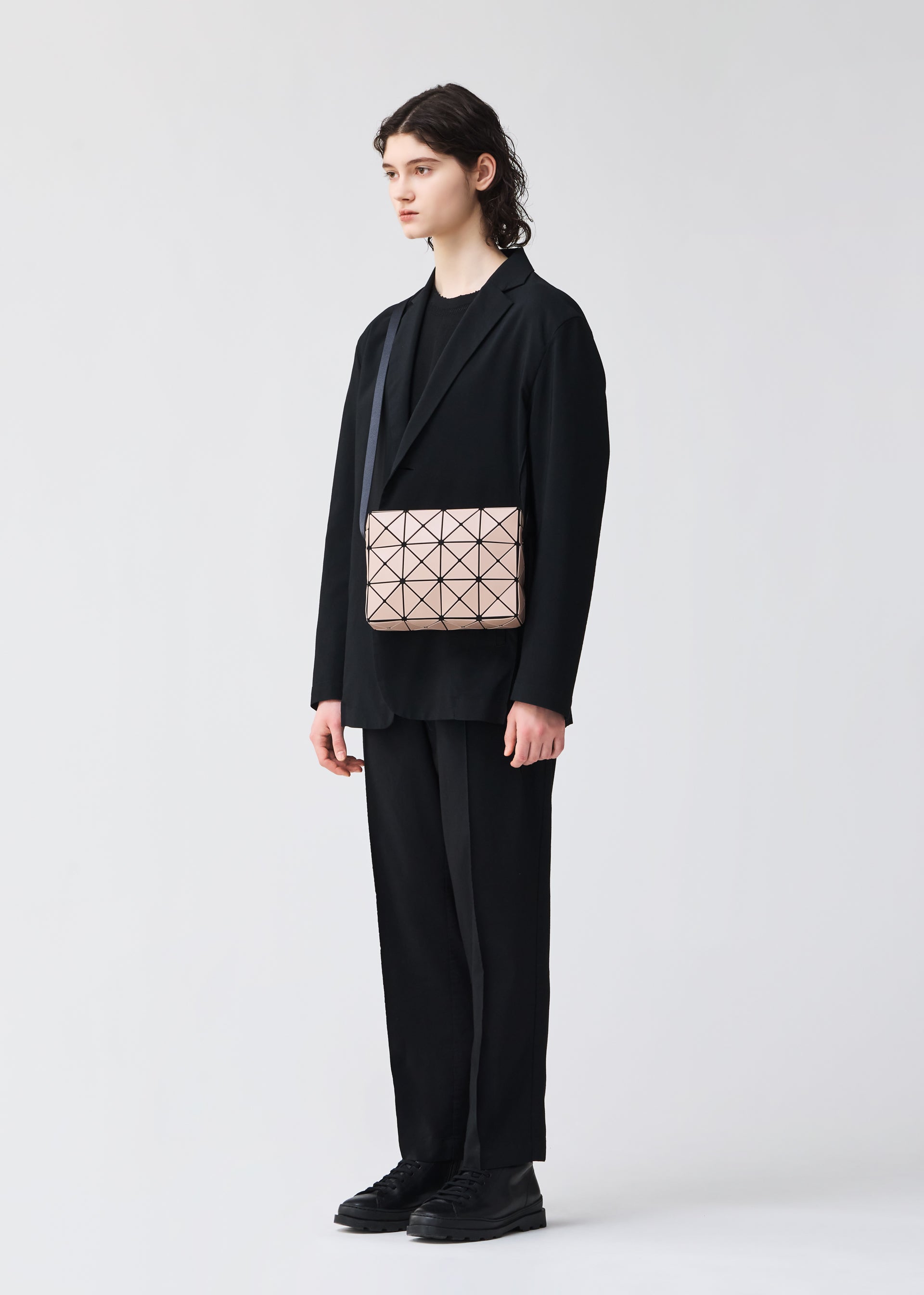 Zoomed view A person with long dark hair in a black suit, T-shirt, and shoes holds the BAO BAO ISSEY MIYAKE CUBOID—a beige minimalist bag with geometric patterns and an adjustable strap—against a plain light background.