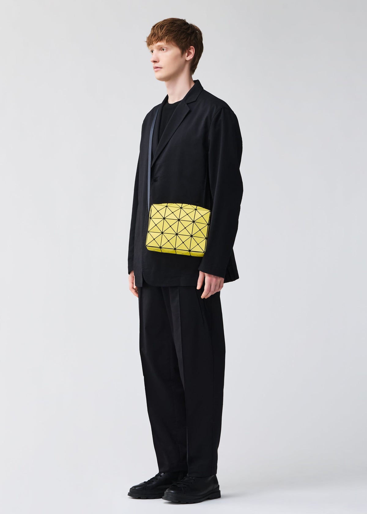 A person with short light brown hair wears a loose black suit and holds the BAO BAO ISSEY MIYAKE CUBOID minimalist yellow shoulder bag under one arm, standing against a plain light background. view 9 of 9