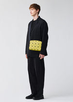 A person in a loose black suit and shoes stands against a plain light background, carrying the BAO BAO ISSEY MIYAKE CUBOID yellow shoulder bag featuring an adjustable strap and bold black triangle pattern across their body.