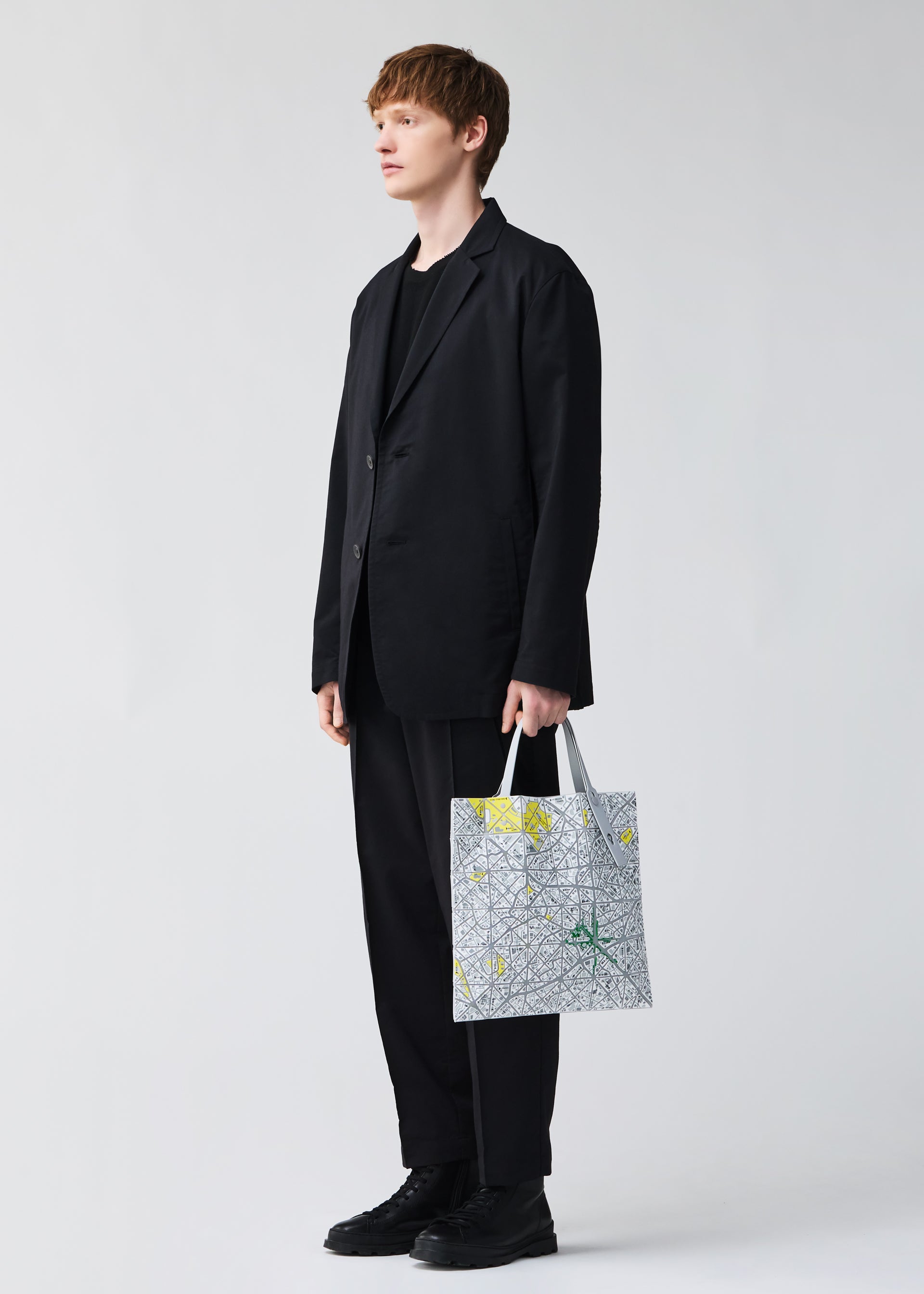 Zoomed view A person with short light brown hair in a black suit and shoes stands holding the BAO BAO ISSEY MIYAKE MAP tote bag featuring a geometric map pattern in green and yellow accents, set against a plain light background.