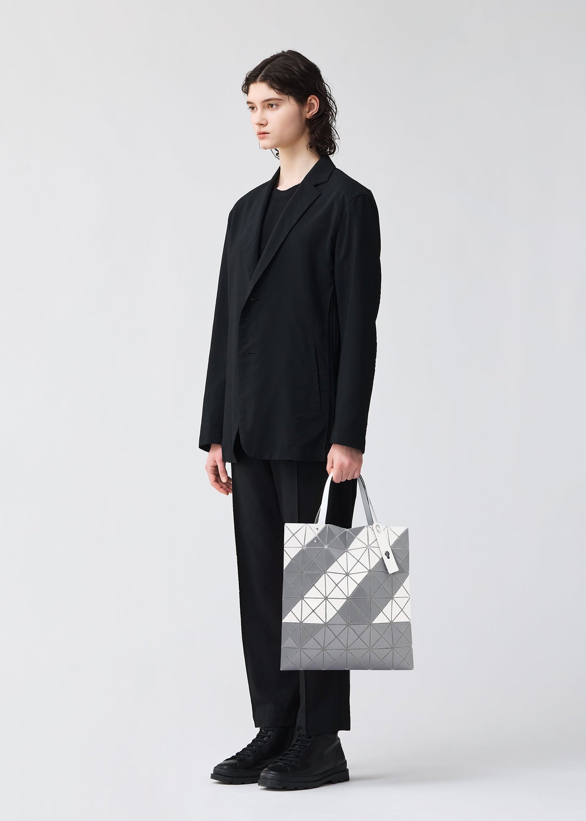 A person with shoulder-length dark hair, wearing a black suit and shoes, stands against a plain white background holding the SIGN tote bag by BAO BAO ISSEY MIYAKE in geometric silver and gray with a zippered interior pocket. view 6 of 6