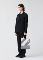 A person with shoulder-length dark hair, wearing a black suit and shoes, stands against a plain white background holding the SIGN tote bag by BAO BAO ISSEY MIYAKE in geometric silver and gray with a zippered interior pocket.