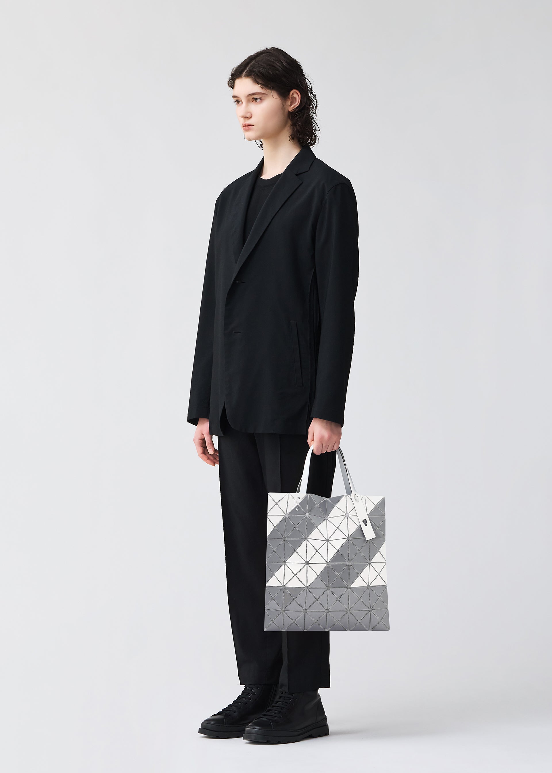 Zoomed view A person with shoulder-length dark hair, wearing a black suit and shoes, stands against a plain white background holding the SIGN tote bag by BAO BAO ISSEY MIYAKE in geometric silver and gray with a zippered interior pocket.
