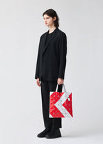 A person with shoulder-length hair in a black suit and shoes stands against a plain light background, holding the BAO BAO ISSEY MIYAKE SIGN tote bag in geometric red, white, and gray—ideal for A4 documents or a 13-inch laptop.