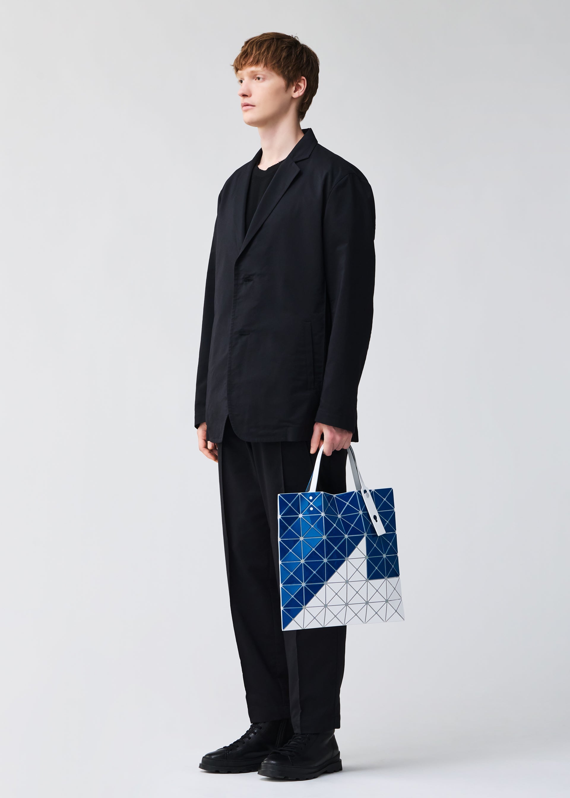 Zoomed view A person in a black suit and shoes holds the SIGN tote bag by BAO BAO ISSEY MIYAKE, featuring a geometric blue and white pattern, ideal for A4 documents or a 13-inch laptop, against a plain light background.