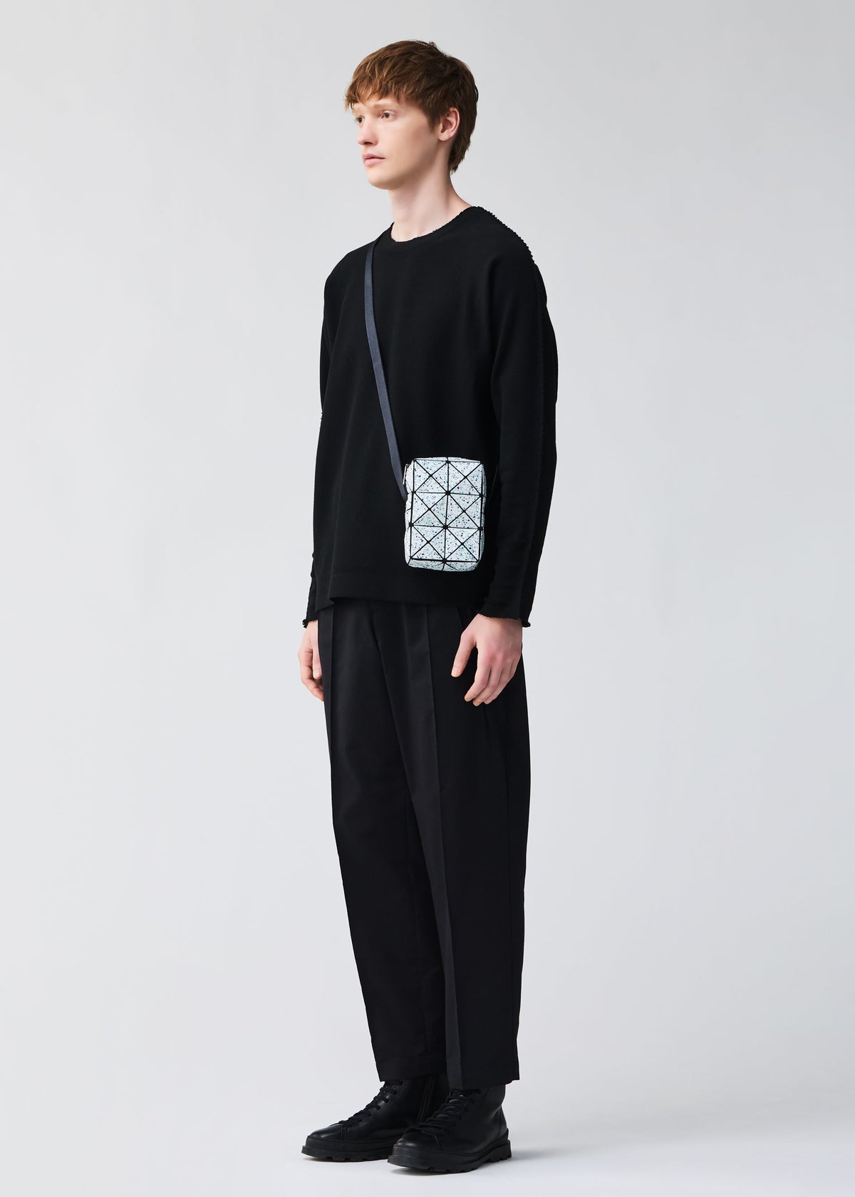 A person stands against a plain light background, styled in all-black attire and carrying the CITY shoulder bag by BAO BAO ISSEY MIYAKE—a small white bag with an adjustable strap and geometric pattern. view 9 of 9