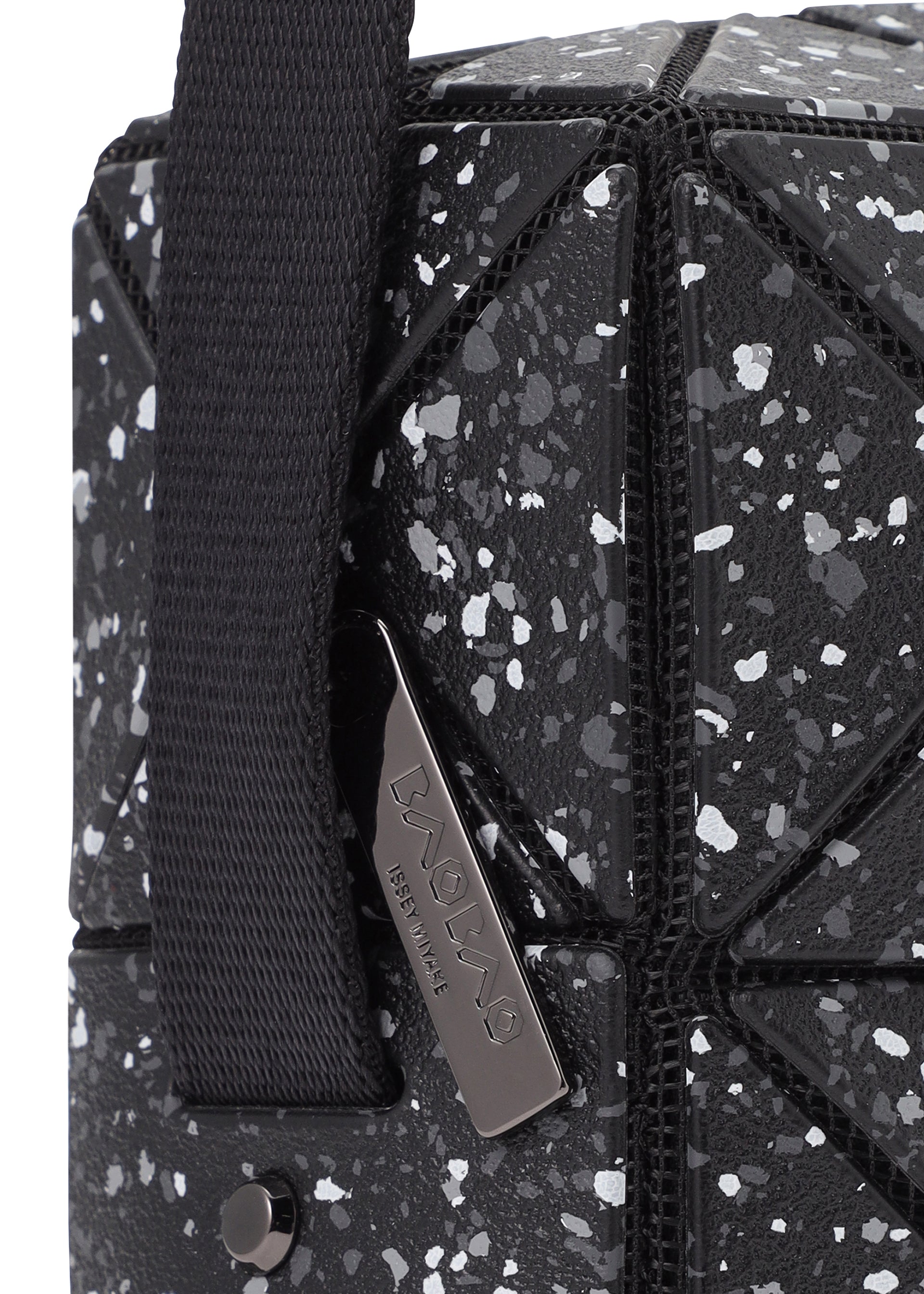 Zoomed view Close-up of the CITY shoulder bag by BAO BAO ISSEY MIYAKE, featuring a black geometric pattern with white speckles, matte leather-like finish, adjustable strap, and a metal tag engraved with the brand name.