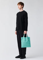 A person stands against a light gray background holding the BAO BAO ISSEY MIYAKE MINIATURE tote, featuring turquoise geometric patterns and large metal hardware, while dressed in black attire.