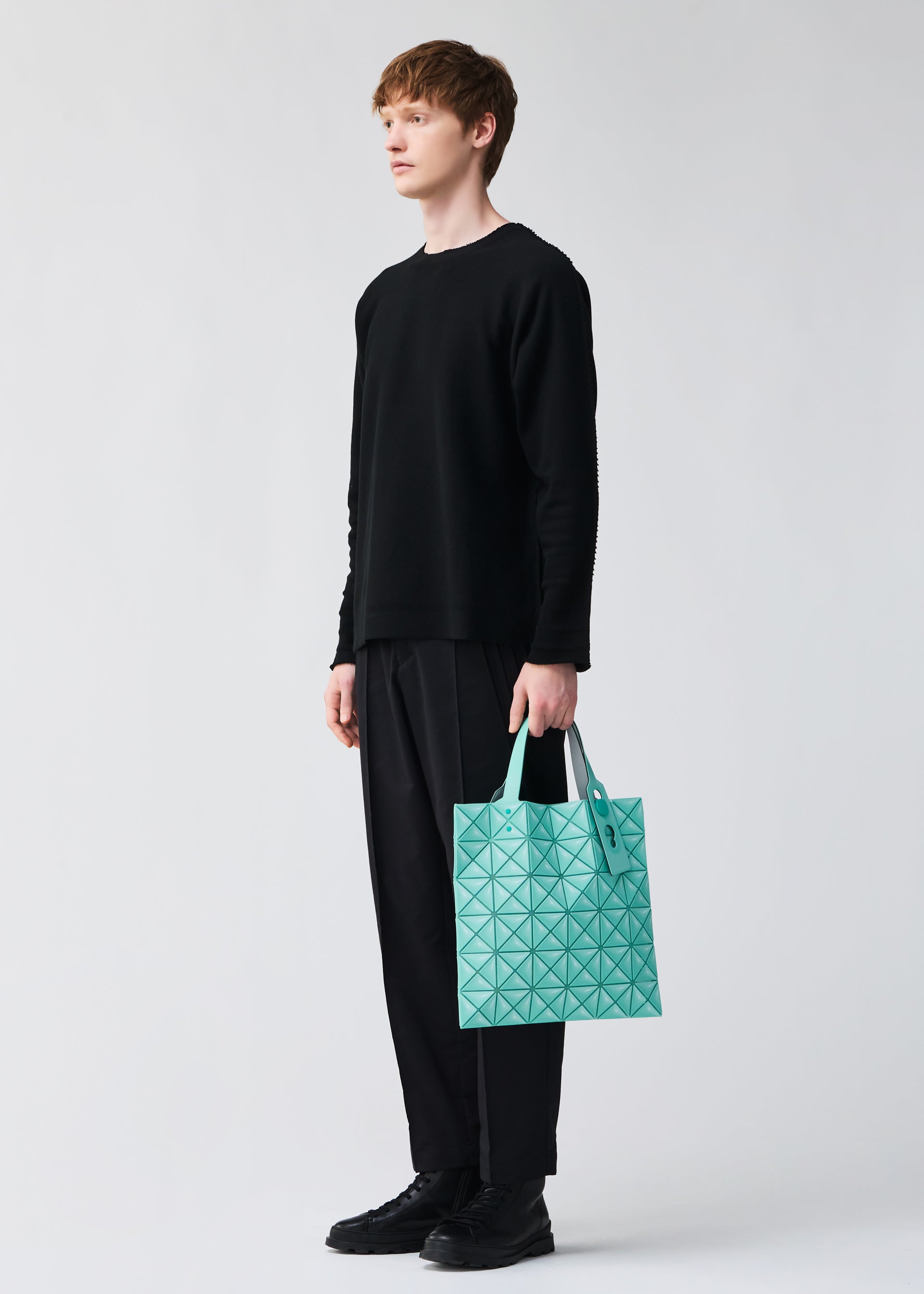 Zoomed view A person stands against a light gray background holding the BAO BAO ISSEY MIYAKE MINIATURE tote, featuring turquoise geometric patterns and large metal hardware, while dressed in black attire.
