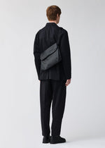 A person dressed in black stands with their back to the camera, carrying the BAO BAO ISSEY MIYAKE FROGGY shoulder bag featuring a black geometric pattern and adjustable straps, set against a plain light background.