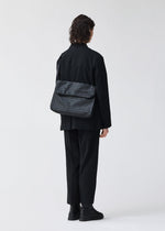 A person with shoulder-length dark hair, dressed in black clothes and shoes, stands facing away while carrying the BAO BAO ISSEY MIYAKE FROGGY—a large shoulder bag featuring adjustable straps and a distinctive patterned design. The bag’s spacious capacity is clearly visible against a plain, light-colored background.