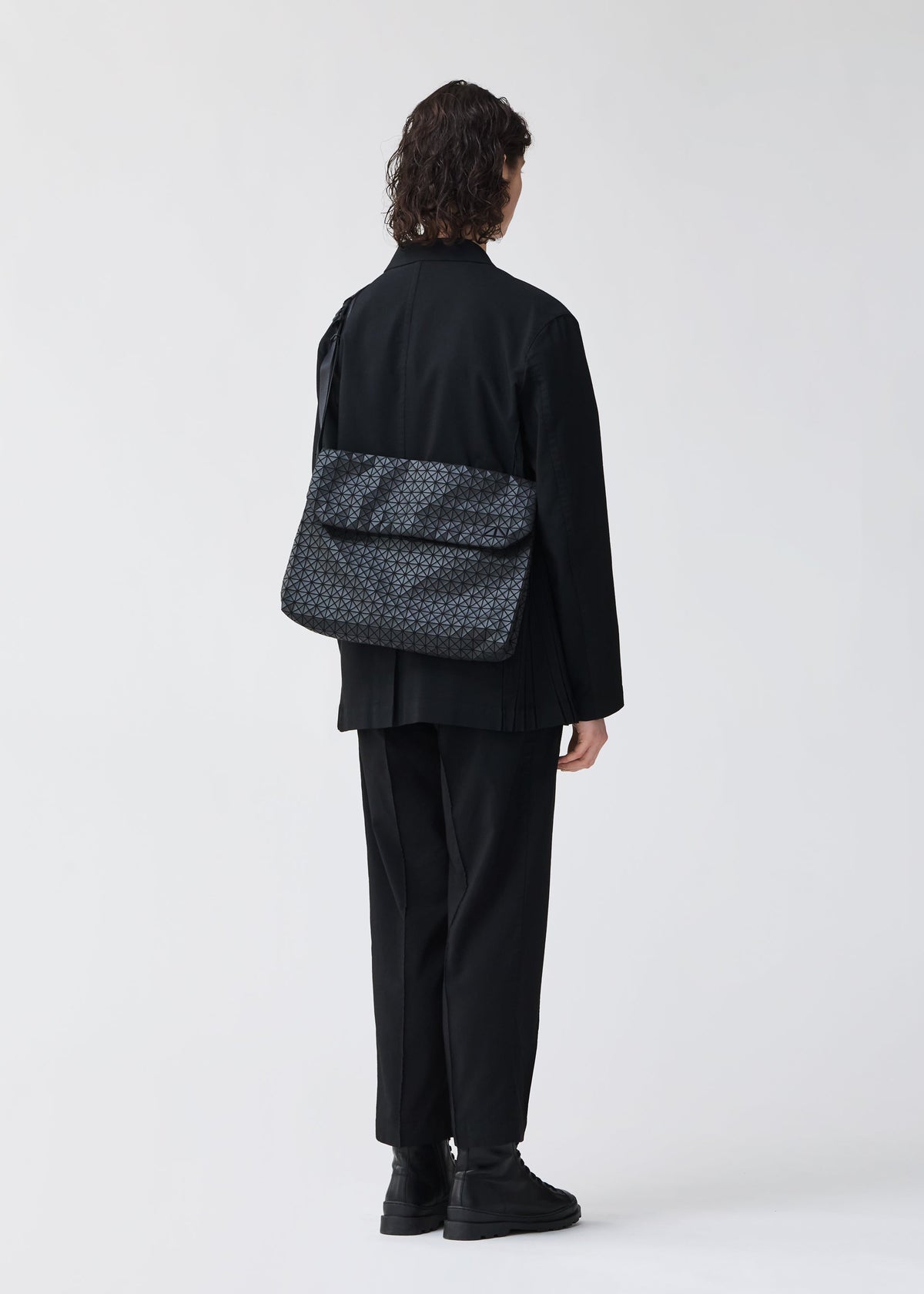 Zoomed view A person with shoulder-length dark hair, dressed in black clothes and shoes, stands facing away while carrying the BAO BAO ISSEY MIYAKE FROGGY—a large shoulder bag featuring adjustable straps and a distinctive patterned design. The bag’s spacious capacity is clearly visible against a plain, light-colored background.