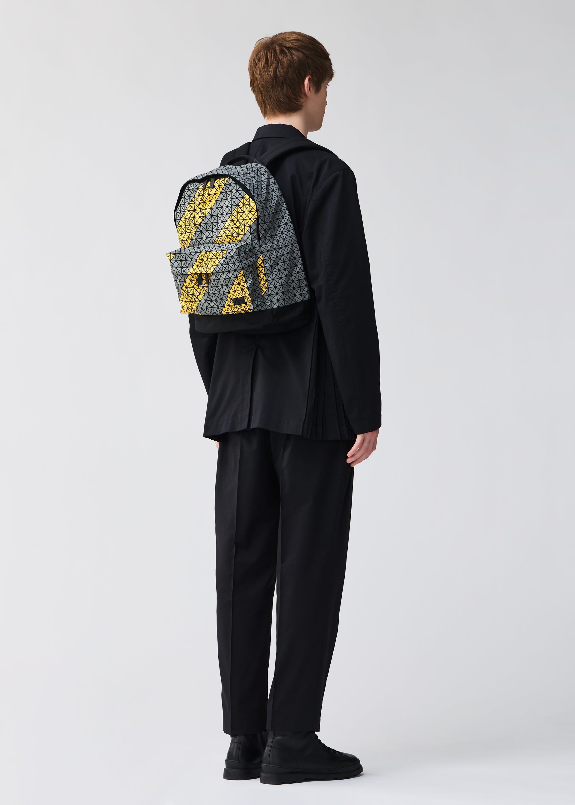 Zoomed view A person with short brown hair, in a loose black outfit and black shoes, stands facing away wearing a BAO BAO ISSEY MIYAKE DAYPACK AUTOBAHN with a gray and yellow pattern. The background is plain and light.