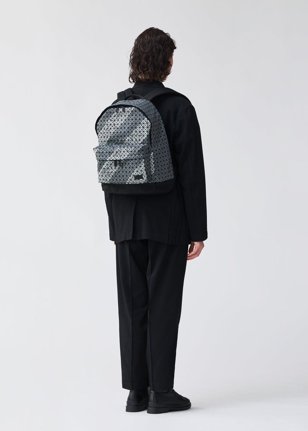 A person with shoulder-length curly hair, in loose black clothes and shoes, stands facing away from the camera wearing a BAO BAO ISSEY MIYAKE DAYPACK AUTOBAHN backpack against a plain white background. view 7 of 7