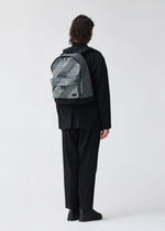 A person with shoulder-length curly hair, in loose black clothes and shoes, stands facing away from the camera wearing a BAO BAO ISSEY MIYAKE DAYPACK AUTOBAHN backpack against a plain white background.