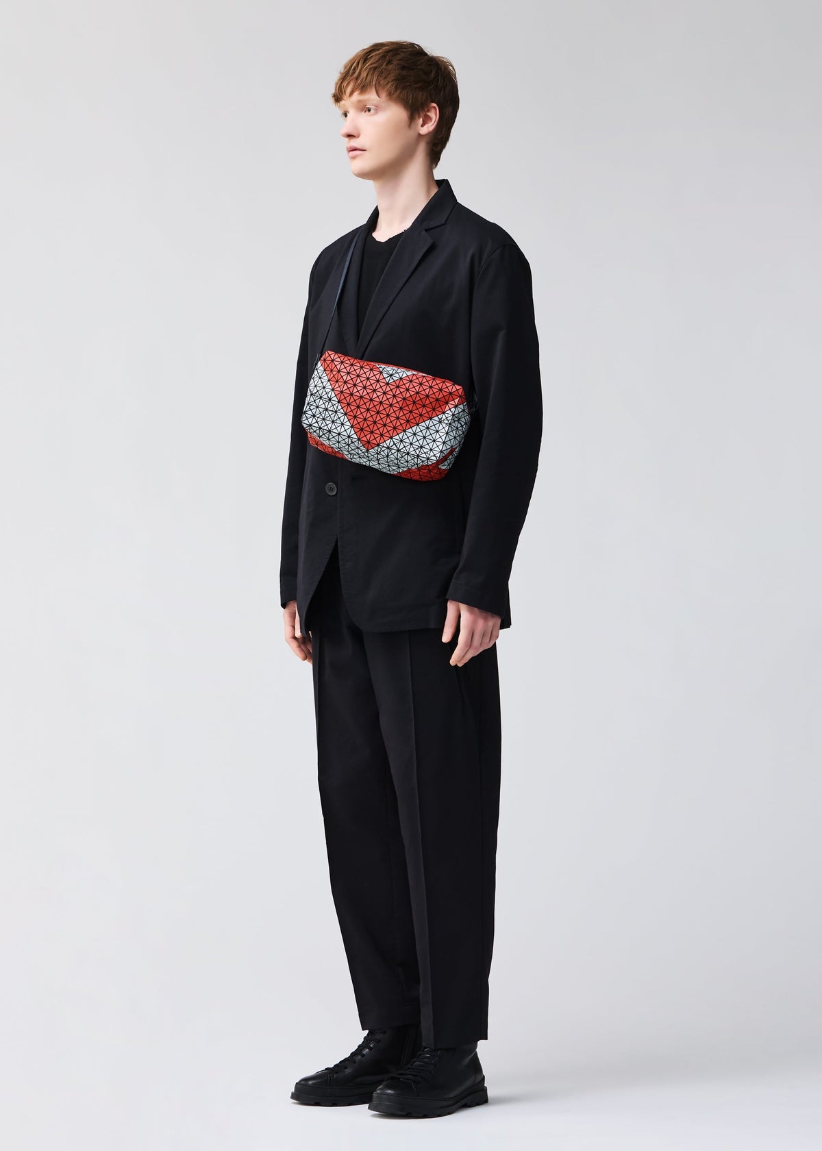 A person stands against a plain background, wearing a loose black suit, black shoes, and a BAO BAO ISSEY MIYAKE SADDLE BAG AUTOBAHN with a bold red and white pattern slung across their chest. view 5 of 6