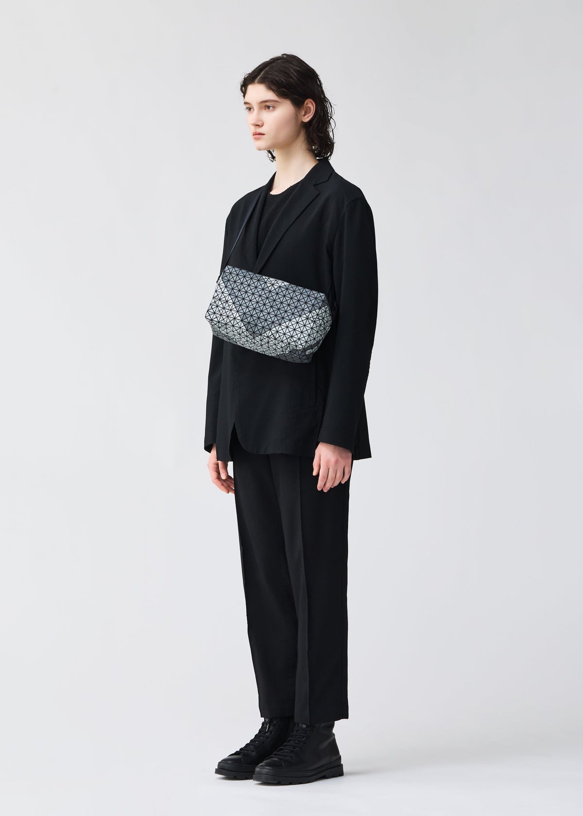 A person with shoulder-length dark hair wears a loose black suit, black shirt, and black boots, standing against a plain light background. They have a BAO BAO ISSEY MIYAKE SADDLE BAG AUTOBAHN crossbody bag across their chest. view 6 of 6