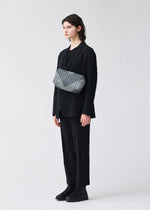 A person with shoulder-length dark hair wears a loose black suit, black shirt, and black boots, standing against a plain light background. They have a BAO BAO ISSEY MIYAKE SADDLE BAG AUTOBAHN crossbody bag across their chest.