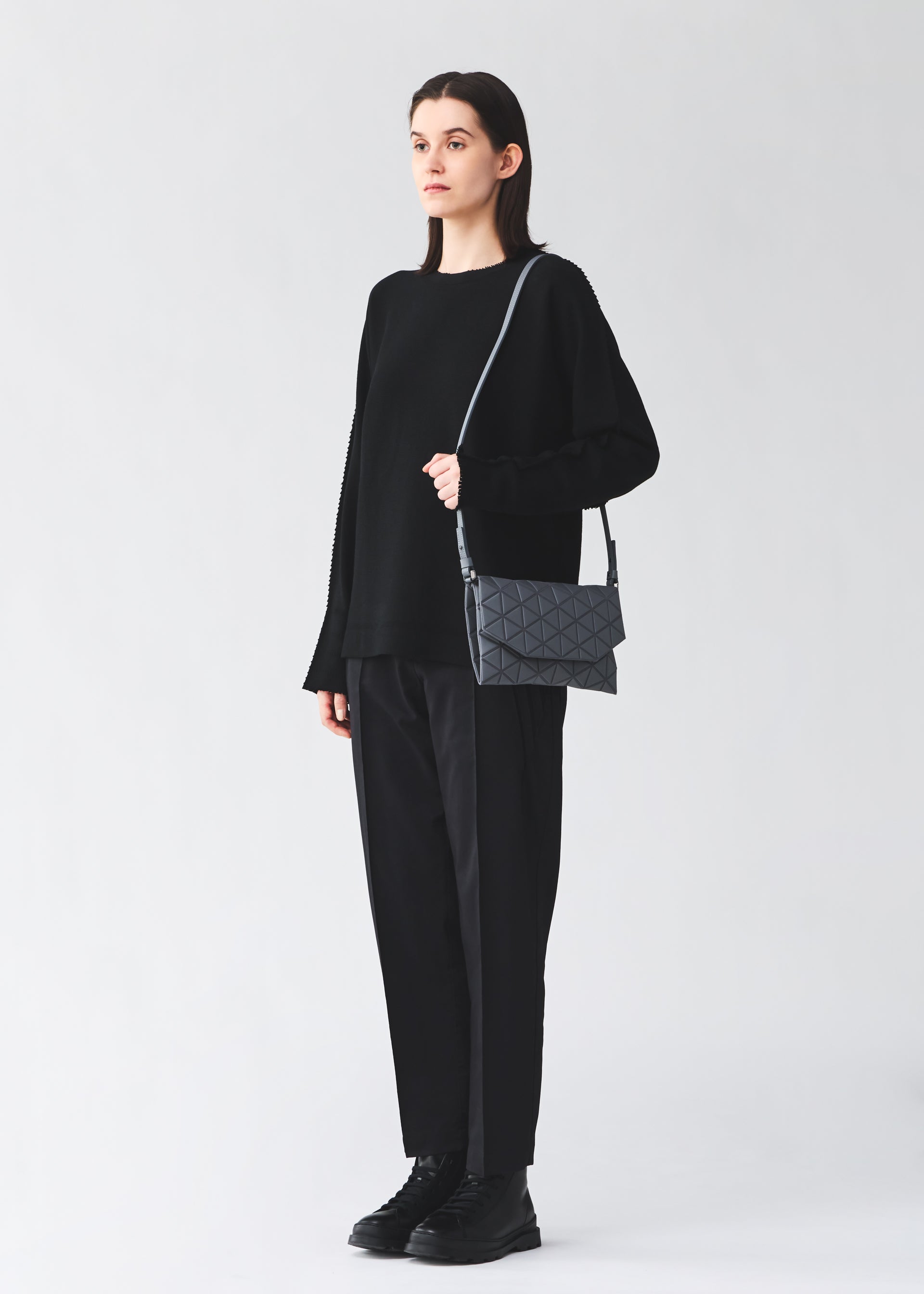 Zoomed view A person wearing a loose black sweater, black pants, and black boots stands holding the BAO BAO ISSEY MIYAKE TONNEAU MATTE shoulder bag—a gray barrel-shaped bag with a geometric pattern—against a plain light background.