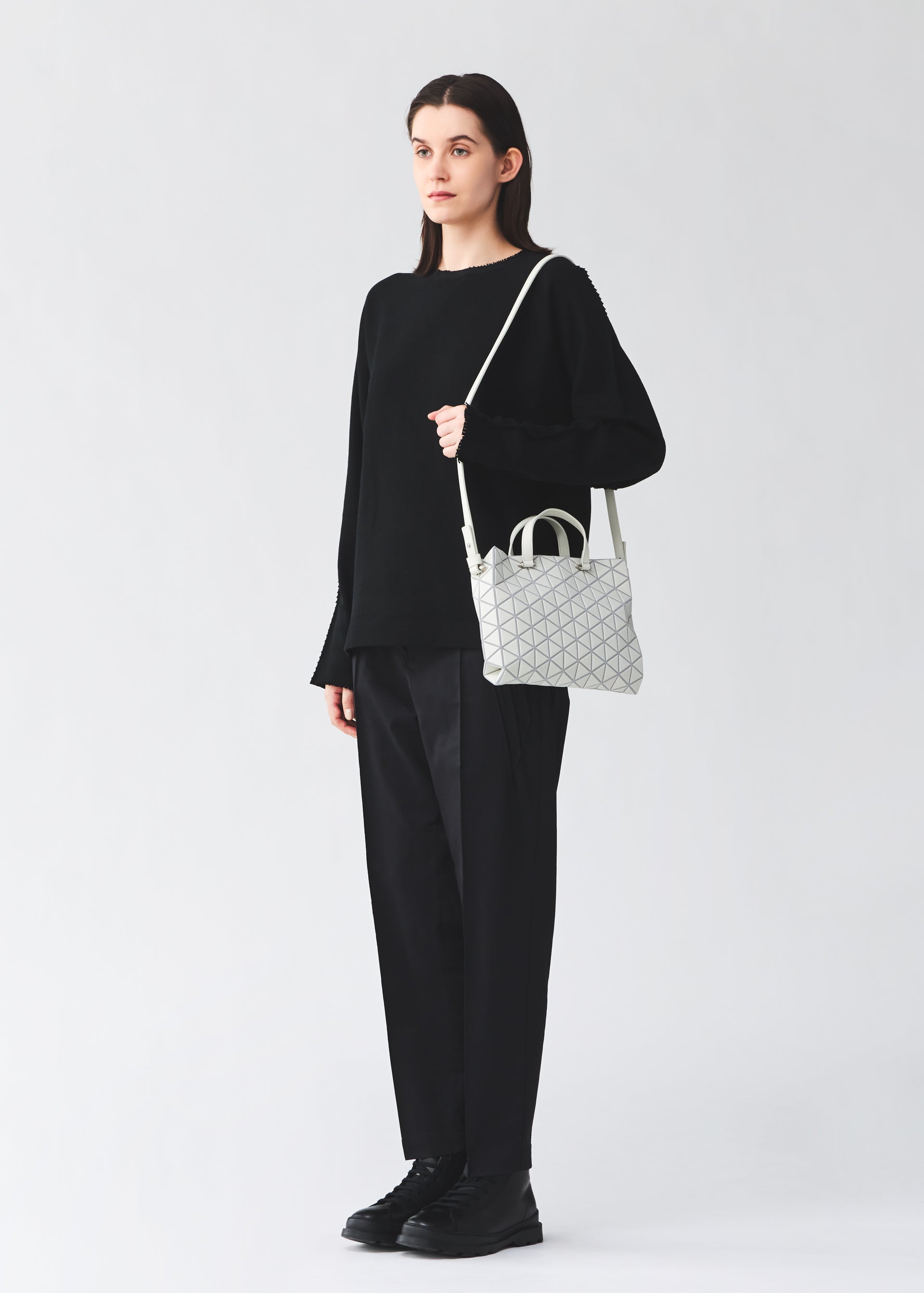Zoomed view A person dressed in a black sweater, black pants, and black shoes stands holding the BAO BAO ISSEY MIYAKE TONNEAU MATTE mini handbag in white, featuring a leather-like matte finish and geometric pattern. The background is plain and light-colored.