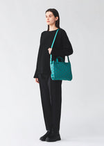 A person dressed in all-black stands against a light background, carrying the BAO BAO ISSEY MIYAKE TONNEAU MATTE handbag with a teal geometric design on their shoulder.