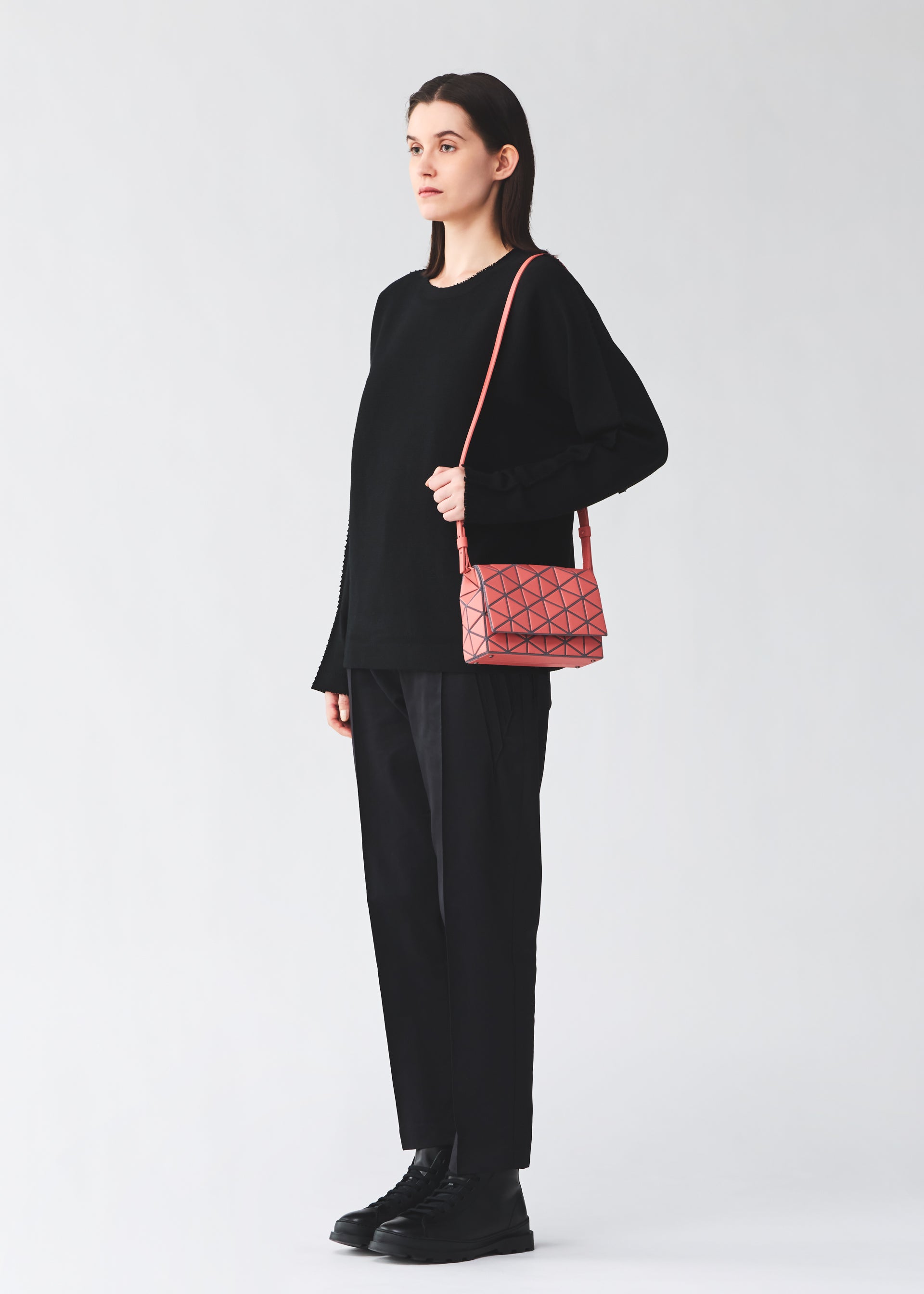 Zoomed view A person stands against a plain background, dressed in a loose black sweater, black pants, and black shoes. They carry the BAO BAO ISSEY MIYAKE FOSSETTE—a small pink cross-body bag featuring a striking geometric pattern and a convenient magnetic closure.