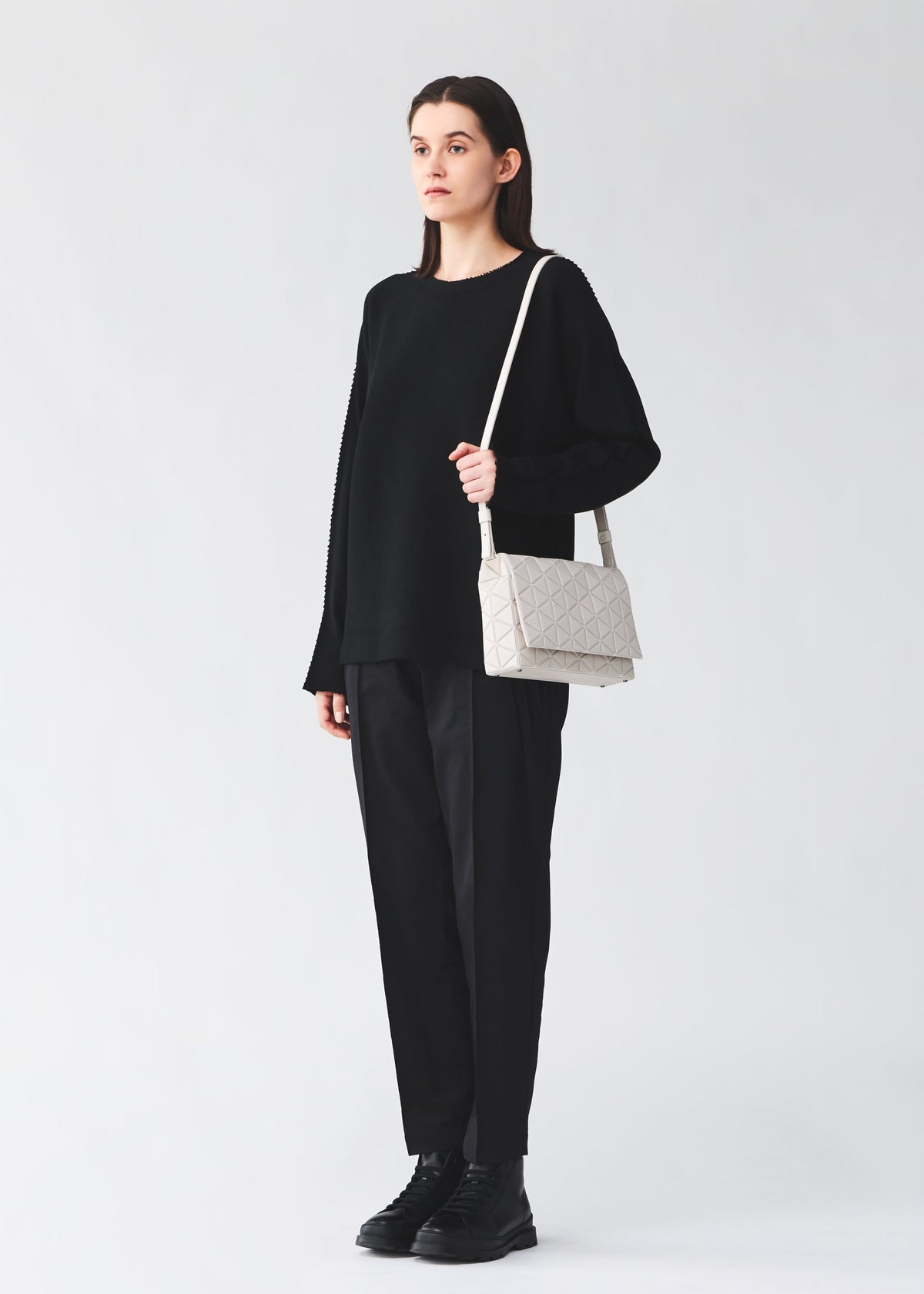 A person stands against a plain background wearing a loose black sweater, black pants, and black boots, holding the BAO BAO ISSEY MIYAKE FOSSETTE cross-body bag, which features a light-colored geometric pattern and a magnetic closure. view 6 of 6