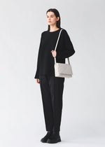 A person stands against a plain background wearing a loose black sweater, black pants, and black boots, holding the BAO BAO ISSEY MIYAKE FOSSETTE cross-body bag, which features a light-colored geometric pattern and a magnetic closure.