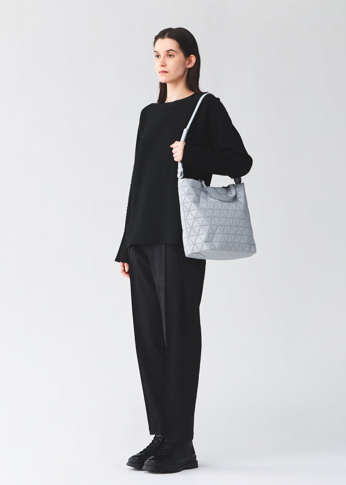 Wearing a loose black sweater, pants, and shoes, a person holds the BAO BAO ISSEY MIYAKE CRYSTAL MATTE handbag—chic with leather straps and perfectly sized for A4 documents—against a plain light background. view 5 of 5
