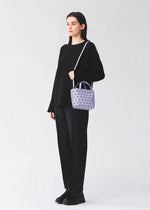 A person stands against a plain light background, wearing all black and carrying the BAO BAO ISSEY MIYAKE CRYSTAL MATTE mini handbag with detachable leather straps and a geometric-patterned lavender design over their shoulder.