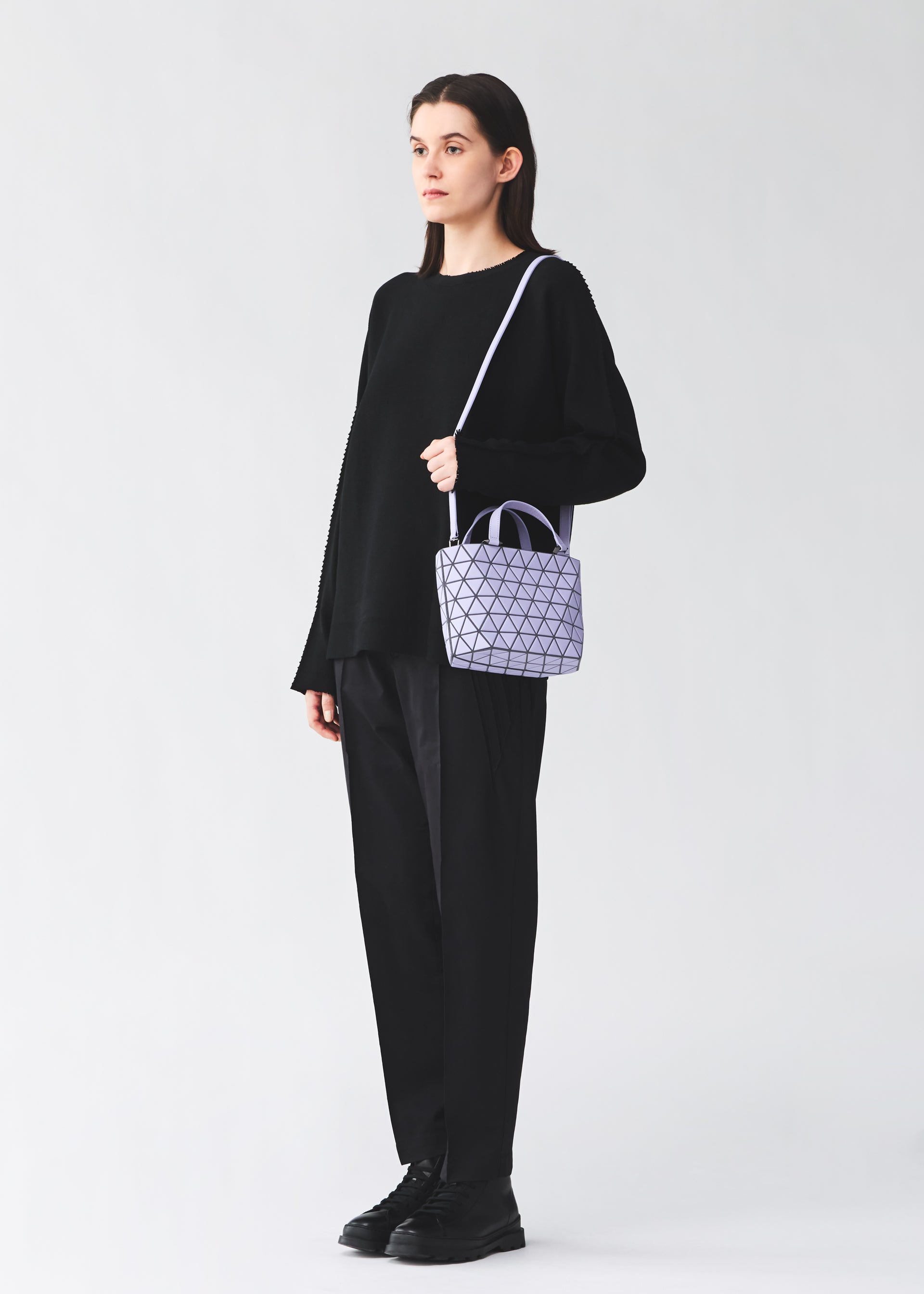 Zoomed view A person stands against a plain light background, wearing all black and carrying the BAO BAO ISSEY MIYAKE CRYSTAL MATTE mini handbag with detachable leather straps and a geometric-patterned lavender design over their shoulder.