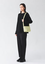 A person with long dark hair stands facing forward in black attire, carrying the BAO BAO ISSEY MIYAKE CRYSTAL MATTE shoulder bag, which features a light yellow color, leather straps, and a geometric pattern against a plain light gray background.
