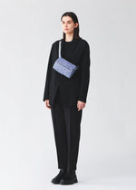 A person in a loose black suit and shoes stands against a white background, carrying the HEXAGON blue geometric-patterned sporty shoulder bag by BAO BAO ISSEY MIYAKE.