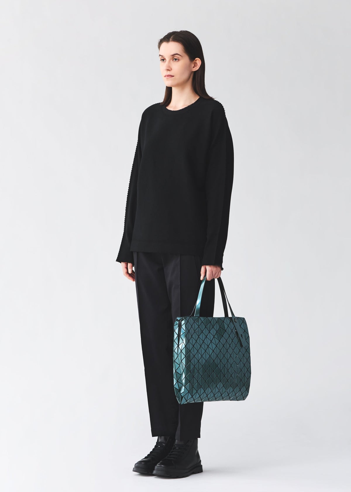 A person stands facing forward against a plain light background, dressed in a loose black sweater, black pants, and black shoes, holding the BAO BAO ISSEY MIYAKE ROW METALLIC handbag—crafted from polarized pearl material with a teal geometric pattern—in their left hand. view 7 of 7