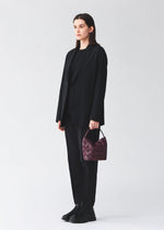 A person in a black suit and boots holds the BAO BAO ISSEY MIYAKE VALLEY, a small, gusseted purple handbag with a geometric pattern, against a plain white background.