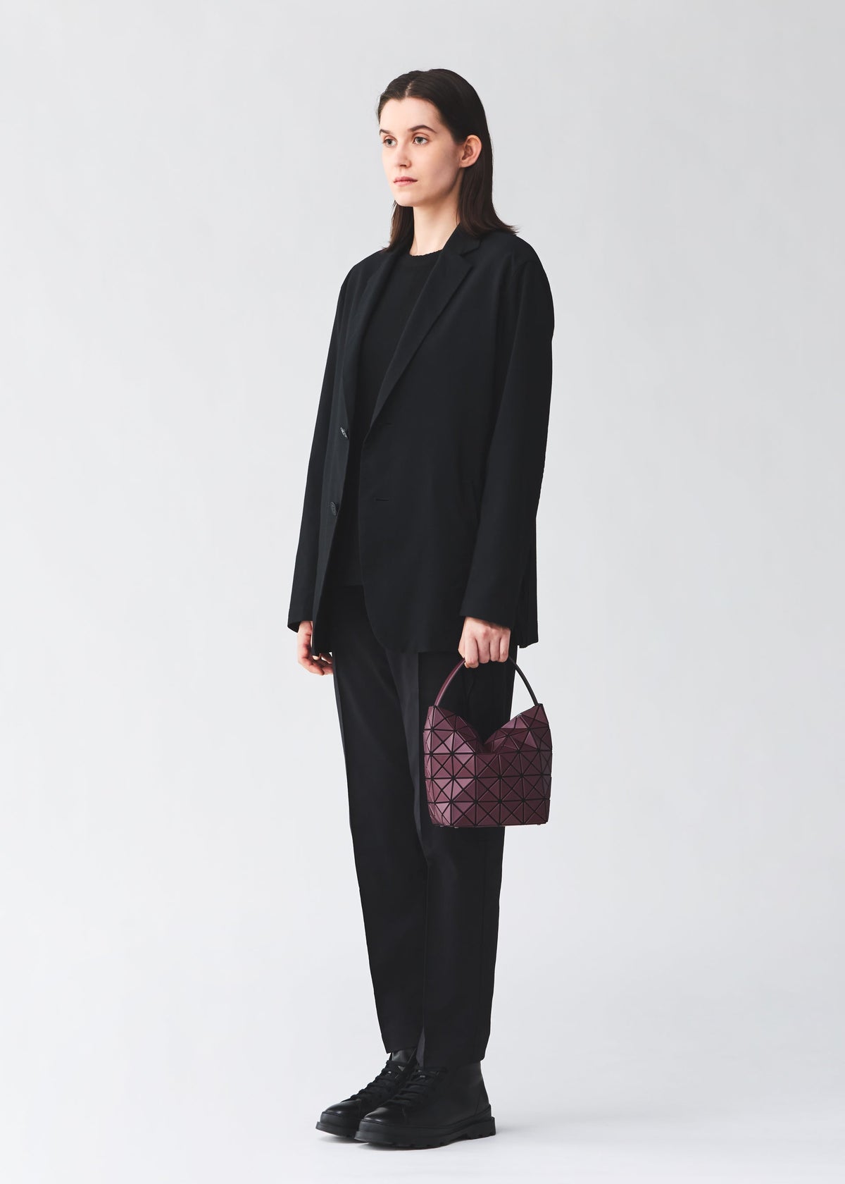 Zoomed view A person in a black suit and boots holds the BAO BAO ISSEY MIYAKE VALLEY, a small, gusseted purple handbag with a geometric pattern, against a plain white background.