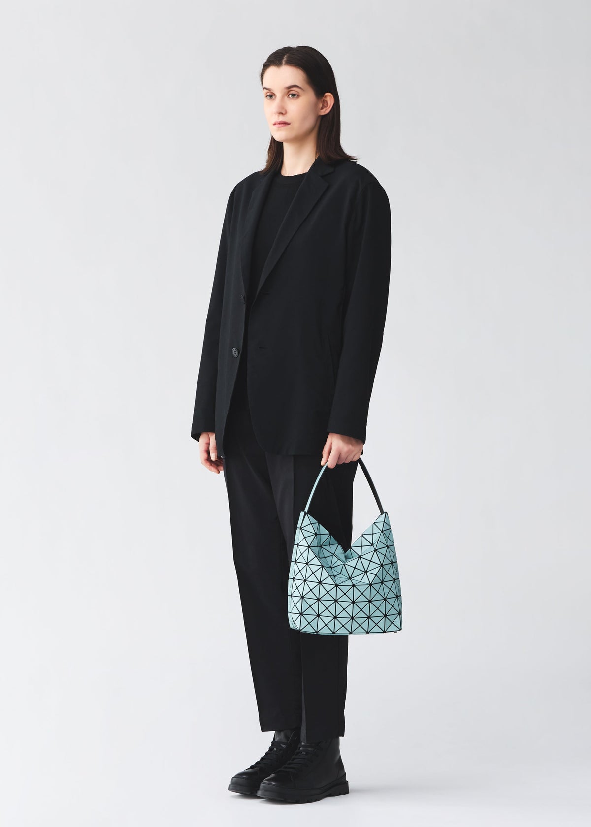 A person in a black suit and boots stands against a plain background, holding the VALLEY handbag by BAO BAO ISSEY MIYAKE—a light blue, self-standing bag with a geometric pattern—in their left hand. view 6 of 6