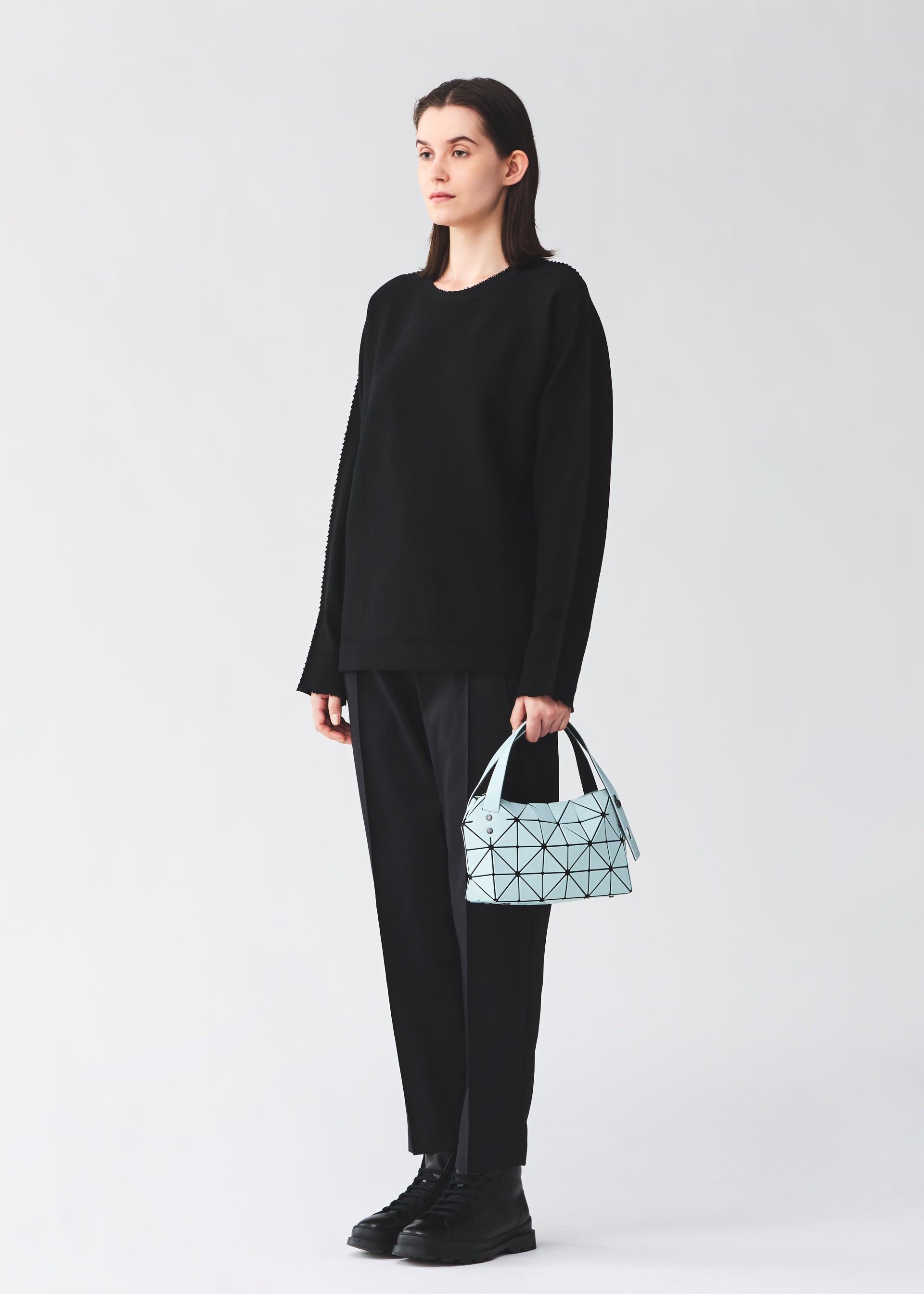 Zoomed view A person with straight brown hair stands against a plain background, wearing black clothes and boots, holding the BAO BAO ISSEY MIYAKE BOSTON mini bag in light blue matte leather-like material with a geometric pattern.