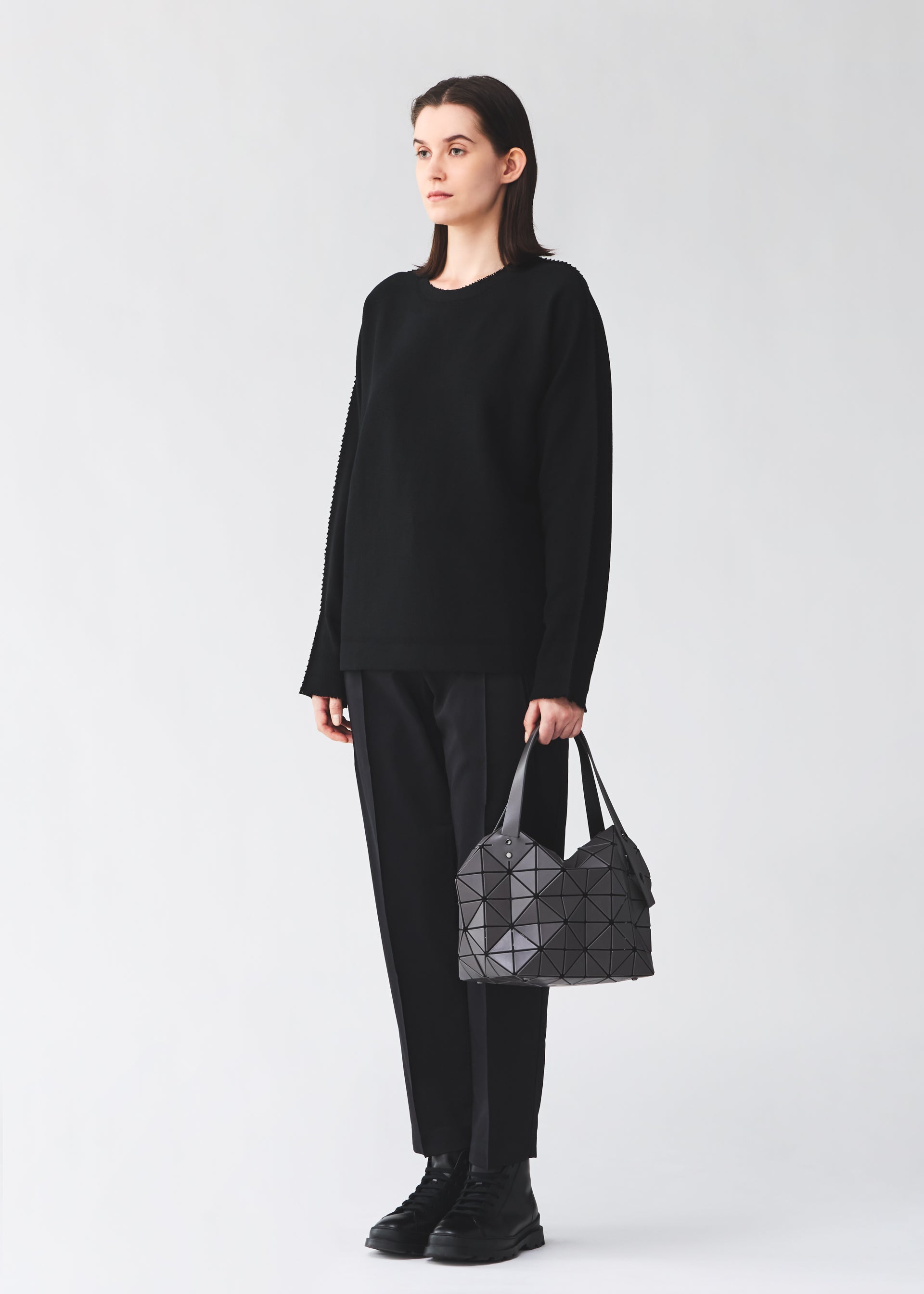 Zoomed view A person with straight brown hair, dressed in a loose black sweater, pants, and boots, stands against a plain light background holding the BAO BAO ISSEY MIYAKE BOSTON bag—a modern gray accessory with ample storage and geometric patterns.