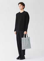 A person dressed in a black sweater, black pants, and black shoes stands holding the BAO BAO ISSEY MIYAKE PRISM GLOSS tote bag in white, its enamel-like finish and geometric design creating a striking contrast against a plain light background.