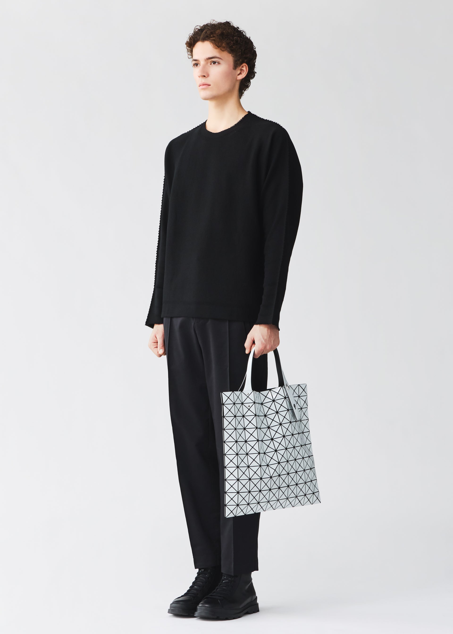 Zoomed view A person dressed in a black sweater, black pants, and black shoes stands holding the BAO BAO ISSEY MIYAKE PRISM GLOSS tote bag in white, its enamel-like finish and geometric design creating a striking contrast against a plain light background.