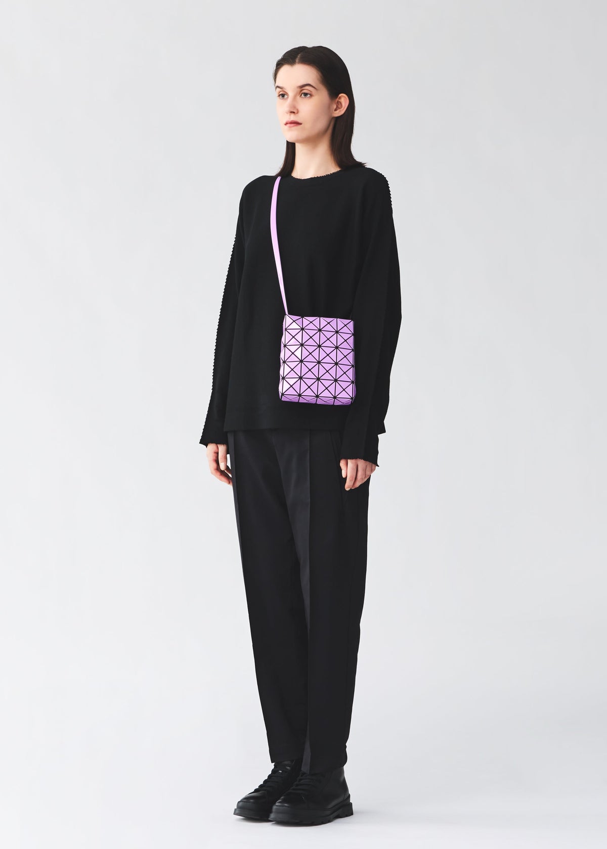A person dressed in all-black clothing and black shoes stands against a plain light background, carrying the BAO BAO ISSEY MIYAKE PRISM GLOSS shoulder bag—a small purple geometric-patterned accessory crafted from an enamel-like material. view 9 of 10