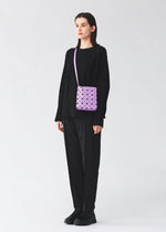 A person dressed in all-black clothing and black shoes stands against a plain light background, carrying the BAO BAO ISSEY MIYAKE PRISM GLOSS shoulder bag—a small purple geometric-patterned accessory crafted from an enamel-like material.