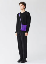 A person dressed in black holds a purple PRISM GLOSS crossbody bag from BAO BAO ISSEY MIYAKE, crafted from an enamel-like material, against a plain light background.