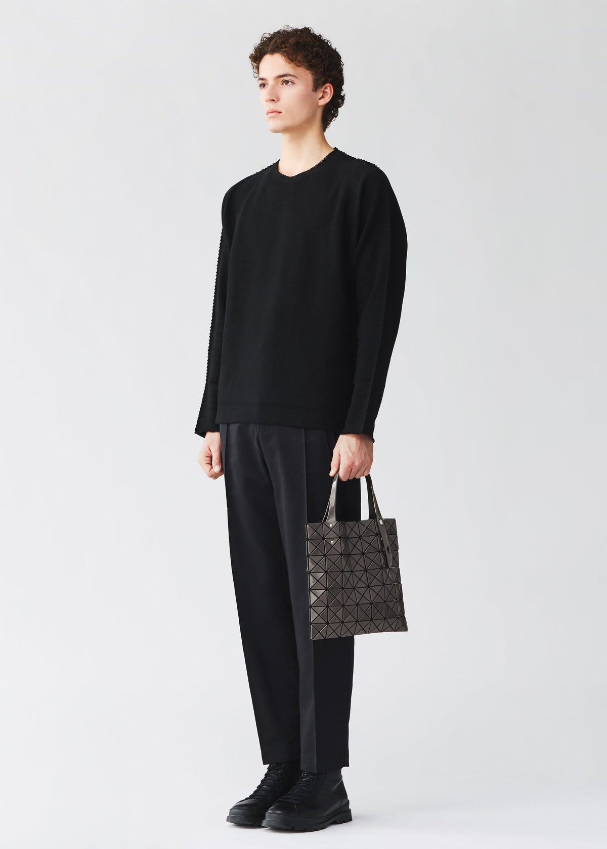 A person stands against a plain light background, dressed in a black sweater, black pants, and black shoes, holding a BAO BAO ISSEY MIYAKE PRISM GLOSS tote bag with a geometric pattern and an enamel-like finish in their left hand. view 10 of 10