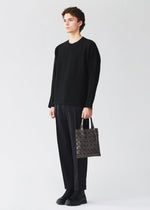 A person stands against a plain light background, dressed in a black sweater, black pants, and black shoes, holding a BAO BAO ISSEY MIYAKE PRISM GLOSS tote bag with a geometric pattern and an enamel-like finish in their left hand.